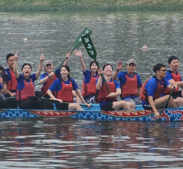 PiA and Fulbright Winner Hannah Semmes on a lake in a dragon boat, racing. The members are on 2 boats that are red and blue with dragon scales, wearing blue shirts, and red life boats. Hannah is in the back, raising a green oar up in the air.
