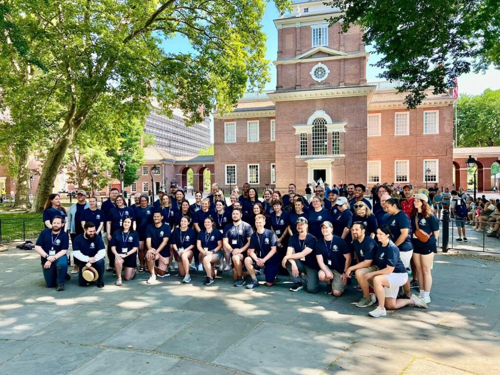 A large group of student kneeling and standing in two rows, taking a group picture together. They are all wearing blue t-shirts, and mixed bottoms with shorts and pants. Behind is a large building with traditional architecture.